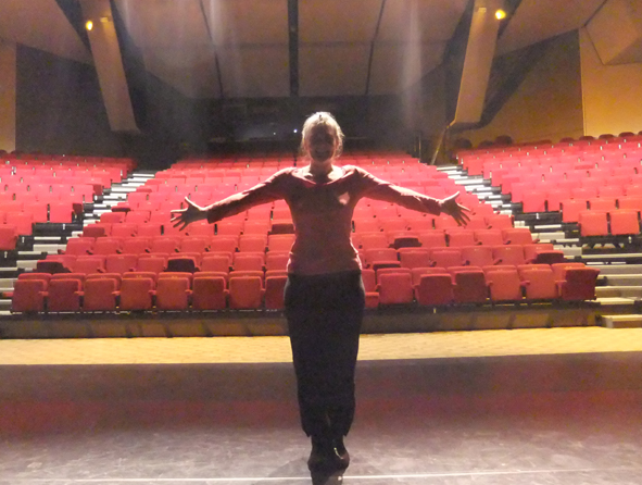 En contre jour on voit Véronique Blot debout, pieds joints et bras tendus
       		devant les gradins rouges de la salle de spectacle de La Flèche dans la Sarthe.