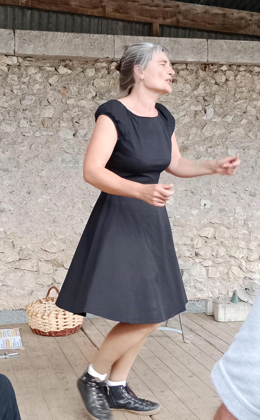 Spectacle Chantons au cœur de la tradition orale avec Véronique Blot, chanteuse.
Crédit photo : Sylvie Chambrin Sur la scène, en extérieur à Villiers sur Loir Loir et Cher, Véronique Blot
en robe et boots noires, est en train de chanter en bougeant les bras et en marchant
sur le plancher en bois. Derrière elle on voit son décor : un panier et une chaise.