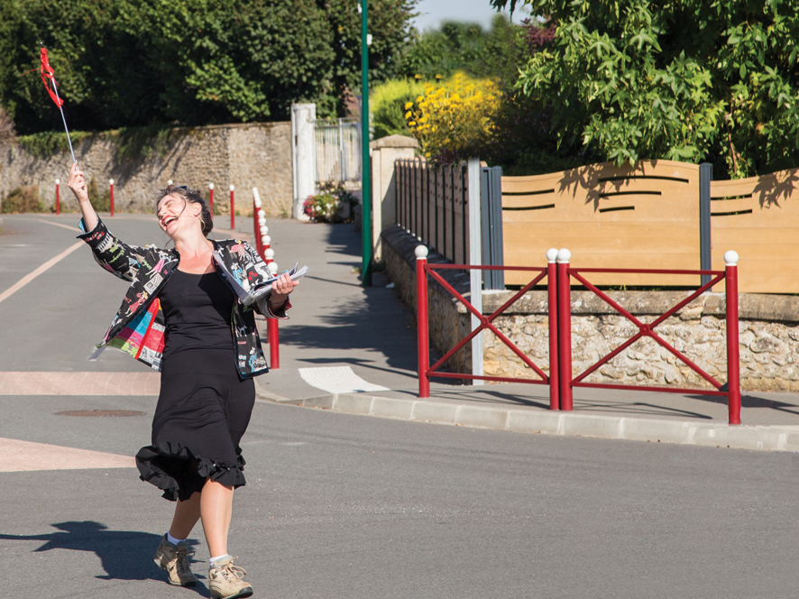 Au milieu d'une rue, Véronique Blot incarne Sylvie Lamour la Super-guide touristique,
        chaussures de randonnée aux pieds, jupe noire au vent, dans un élan en arrière, elle
        éclate de rire et tend en l'air, à bout de bras, une baguette avec fanion rouge pour
        que les touristes la repèrent dans le bourg et la suivent.