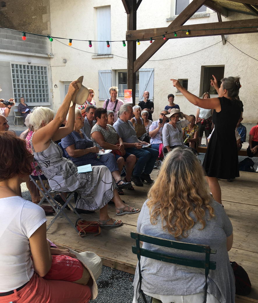 On voit la chanteuse de dos, elle fait face au public assis sous un hangar
       		décoré d'une guirlande de couleur. Véronique a les bras en l'air et certaines personnes
       		du public soulèvent un chapeau. Le public est souriant.